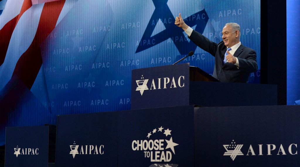 El Premier israelí Netanyahu saludando sonriente desde el podio de oradores en una conferencia de AIPAC, con gesto en señal de aprobación. En el podio mismo, el logo de AIPAC. De fondo, en una pantalla, se ve parte de las banderas de Israel y Estados Unidos.
