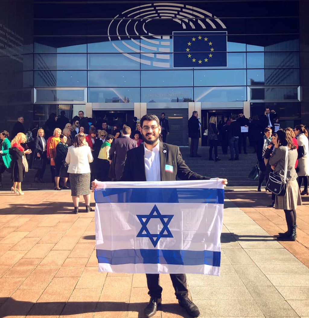 Un joven sonriente, de chaqueta, muestra la bandera de Israel a la entrada del Parlamento Europeo 