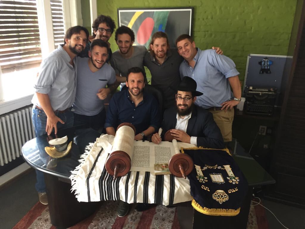 Un grupo de hombres rodea al Rabino Mendy Shemtov, sentado junto a una mesa en la que hay un rollo de la Torá