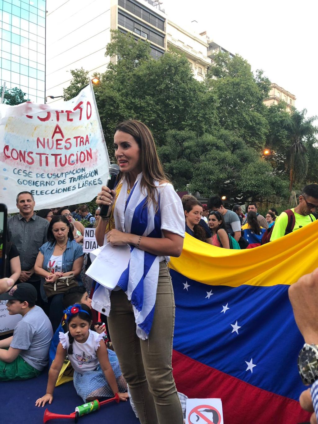 Caro Ache de pie,con la bandera uruguaya al cuello. De fondo, gente sentada en el piso, y una bandera de Venezuela, y carteles.
