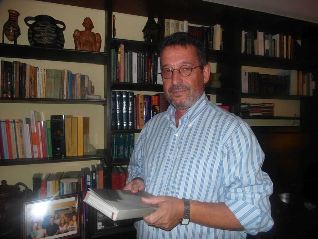 El escritor Fernando Butazzoni, con un libro en sus manos, biblioteca de fondo, en el comedor de su casa
