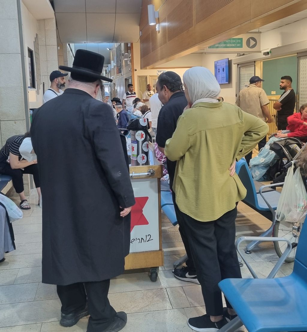 Junto a una estación móvil en la que voluntarios ofrecen comida y bebida a quien quiera recibir mientras espera ser atendido en el hospital Hadassah Ein Karem, una judío religioso y una mujer musulmana aguardan su turno