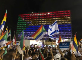manifestación en Tel Aviv, una multitud presente, se ven cabezas más que nada, manos en alto con banderas de Israel y de la comunidad drusa, Y de fondo, una pared, de la municipalidad de Tel Aviv, encendida con luces que forman la bandera de la comunidad 