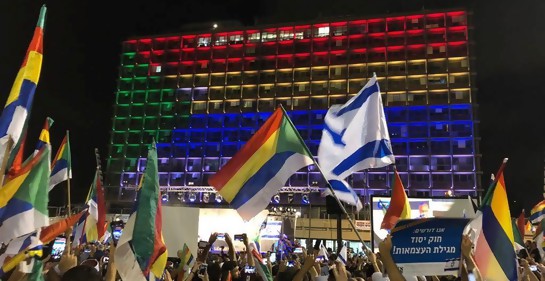 manifestación en Tel Aviv, una multitud presente, se ven cabezas más que nada, manos en alto con banderas de Israel y de la comunidad drusa, Y de fondo, una pared, de la municipalidad de Tel Aviv, encendida con luces que forman la bandera de la comunidad 