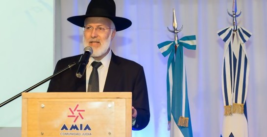 El rabino Davidovich haciendo uso de la palabra en un podio de oradores de AMIA. Vestido de negro, con sombrero negro. De fondo, la bandera de Israel y la de Argenitina.