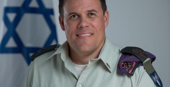 Tte Cnel Jonathan Conricus, de uniforme, boina al hombro, insignias, de fondo la bandera de Israel