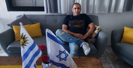 Un hombre sentado en el salón de su casa, de frente dos banderas, de Israel y Uruguay