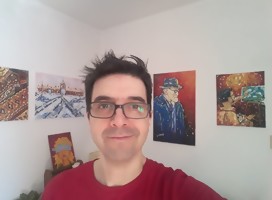 Pablo Solari, de lentes, sonriente, y de fondo sobre una pared blanca, algunos de sus cuadros