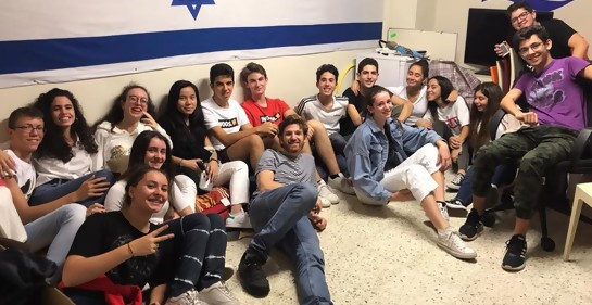 Un grupo de jóvenes sentados en el piso, y detrás, una bandera de Israel