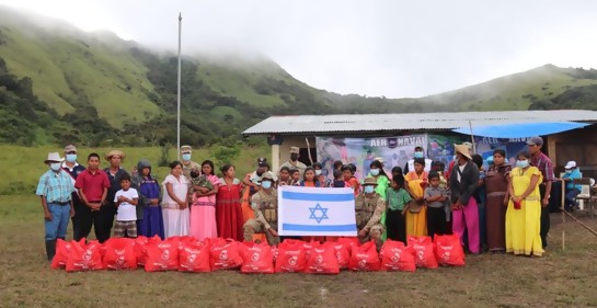 Una fila de personas, con personal del servicio aeronaval, con los paquetes de aliimentos y en el medio la bandera de Israel