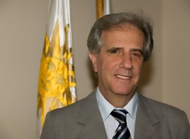 El Dr Vázquez. sonriente. de fondo, la bandera de Uruguay