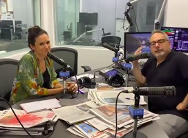 una mujer y un hombre en un estudio radial, micrófono en el medio , mesa llena de diarios