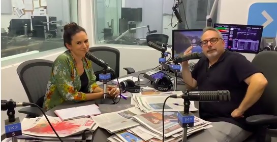 una mujer y un hombre en un estudio radial, micrófono en el medio , mesa llena de diarios